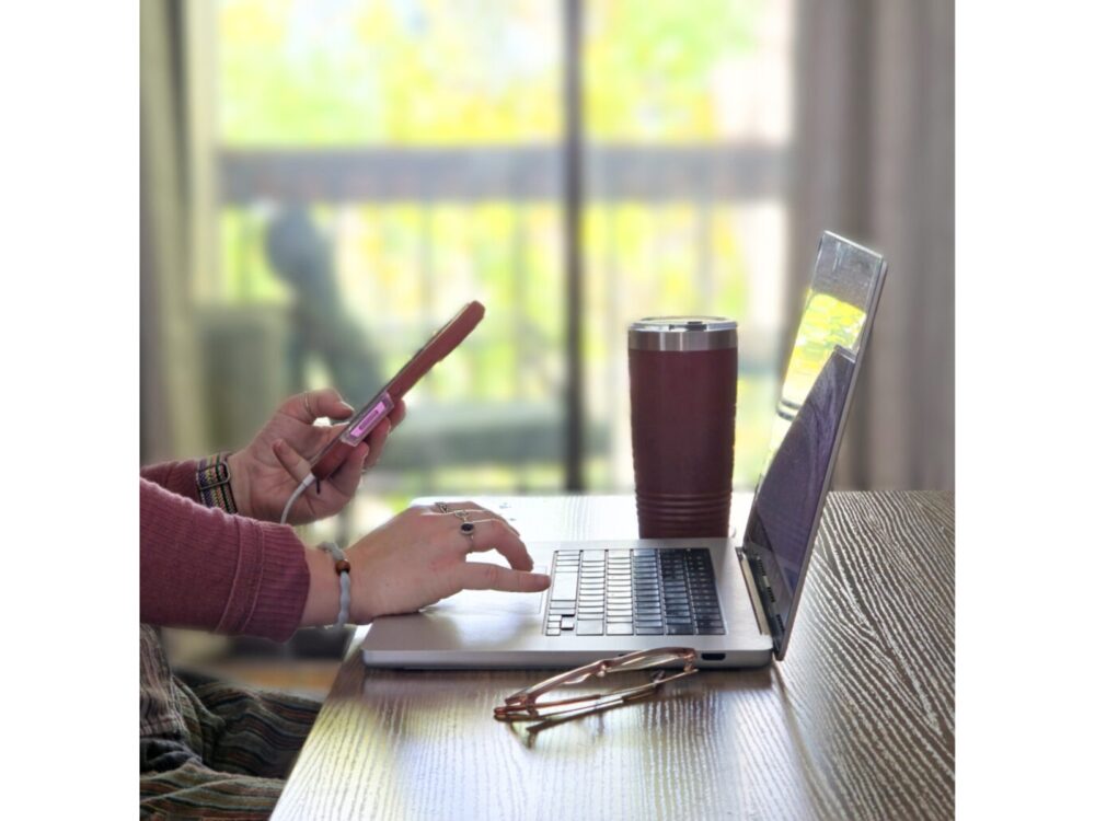 A stock image of a person typing on a computer and holding a cell phone indoors.