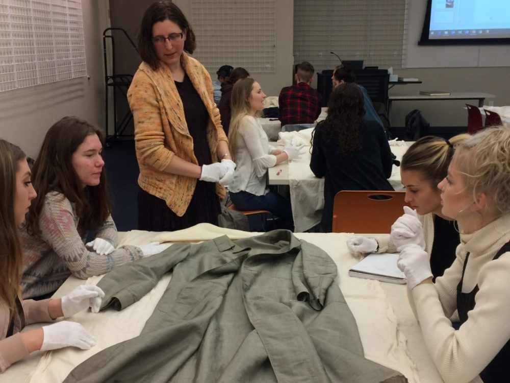 An instructor works with groups of students sitting at tables examining textiles