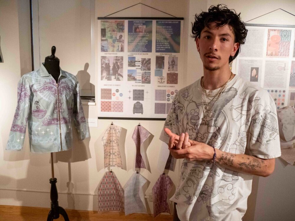 A student standing with a mannequin wearing a jacket and textile designs on the wall in a Gustafson Gallery past exhibit.