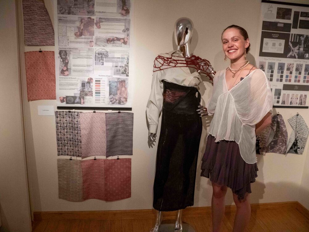 A student standing with a mannequin wearing a dress and textile designs on the wall in a gustafson gallery past exhibit