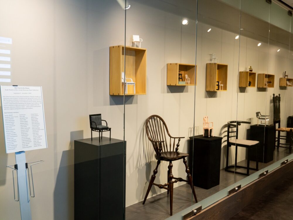 Historic chairs are lined up in a long glass case with small models of them mounted above