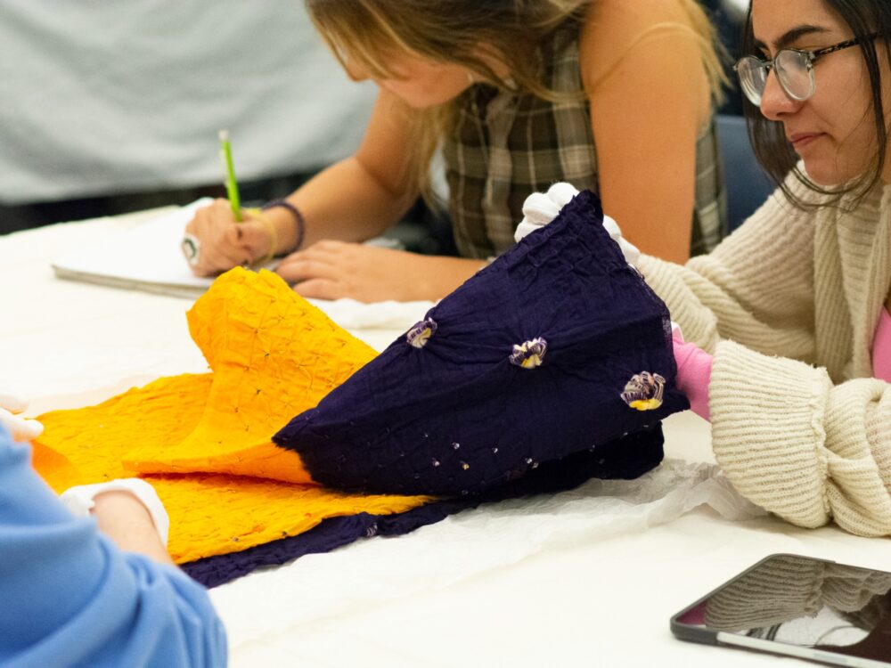 2 students examine a textile up close while wearing gloves while a third student takes notes