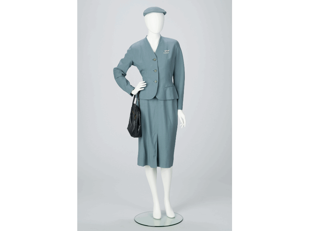 A white museum mannequin wears a robin's egg blue flight attendant's uniform complete with `sugar scoop` style hat and navy blue handbag.