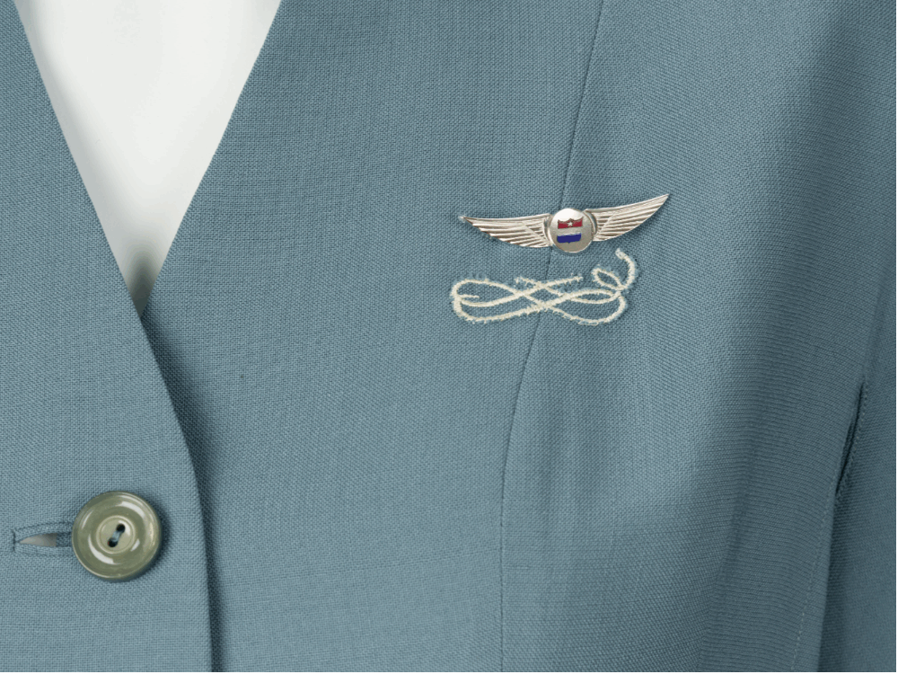 A detail image of a blue flight attendants uniform with a pin on the breast. The pin sits just above a white swirling motif embroidered on the jacket.
