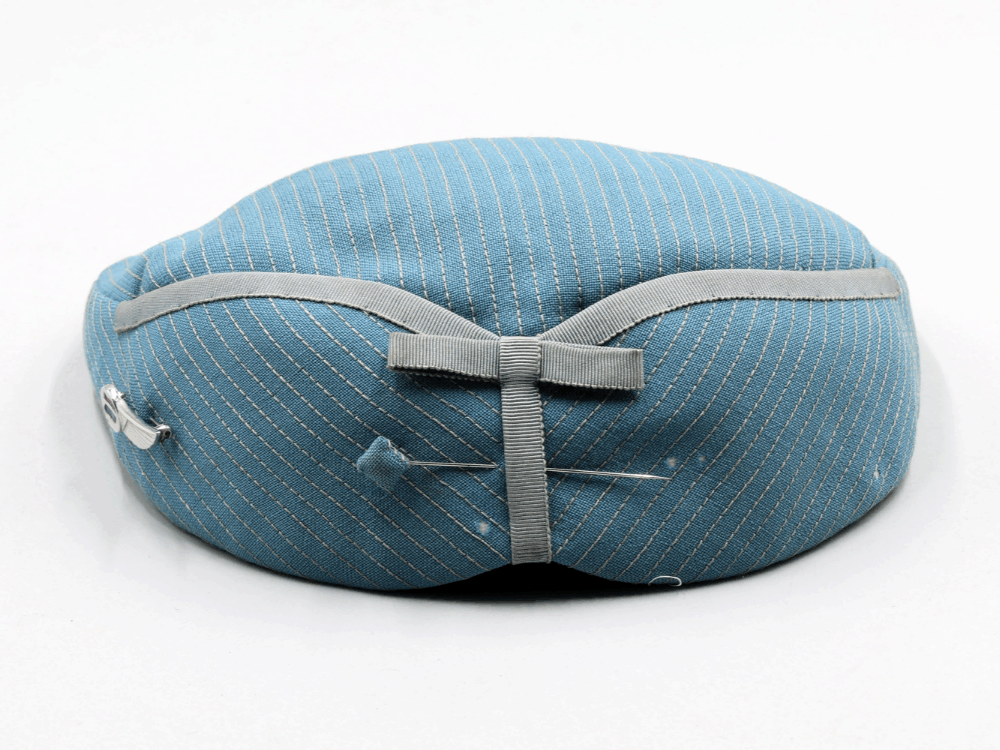 A posterior view of a blue flight attendants hat. The hat features a lin of piping that insinuates a plane's wings. The piping forms a bow at the center back. There is a hat pin with a fabric cover that matches the robin's egg blue of the hat.