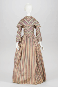 A mannequin wearing a tulle veil over her face is clothed in an ensemble from c. 1850. The bodice and skirt ensemble is made of a striped fabric of cream, purple, and blue. The bodice has a ruffle around the shoulder, and comes to a point at the front.