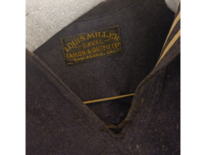 A detail image of the interior tag on a blue winter sailor's blouse. The tag is black with gold lettering and reads: Louis Miller, Naval Tailor & Outtfitter San Pedro, Cal.