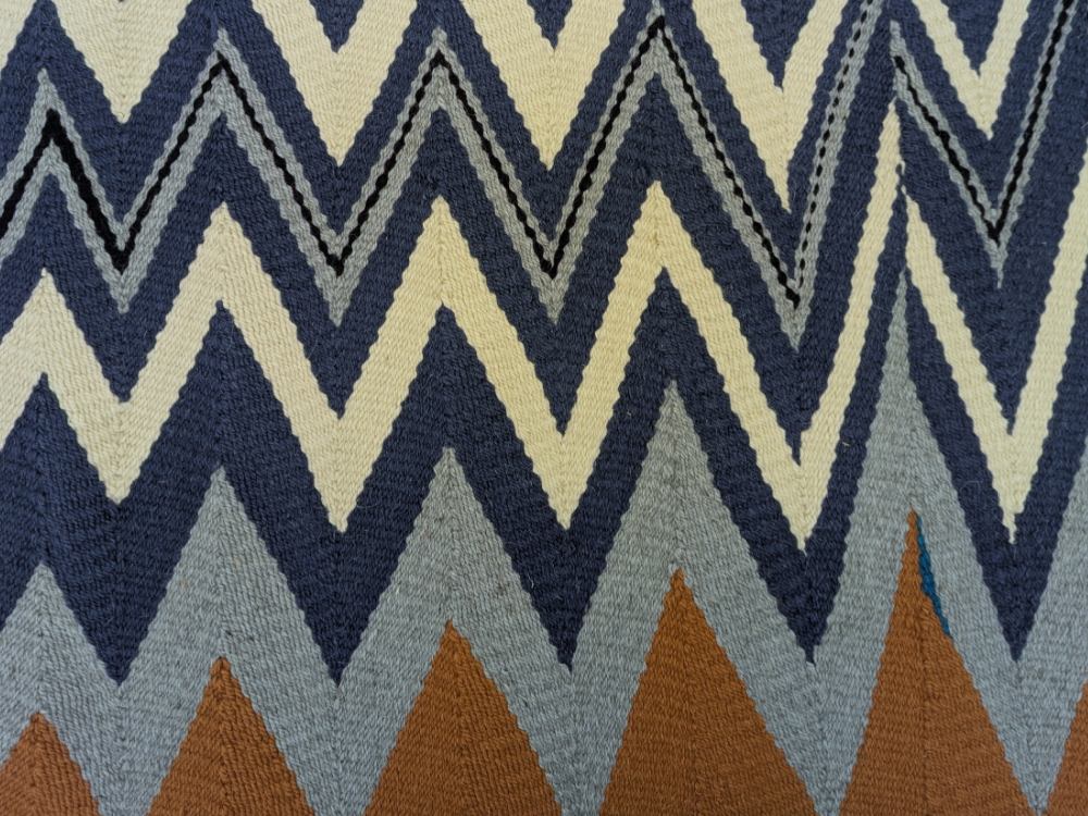 A tapestry woven textiles in navy blue, light blue, and tan. The textile is woven into a chevron pattern.