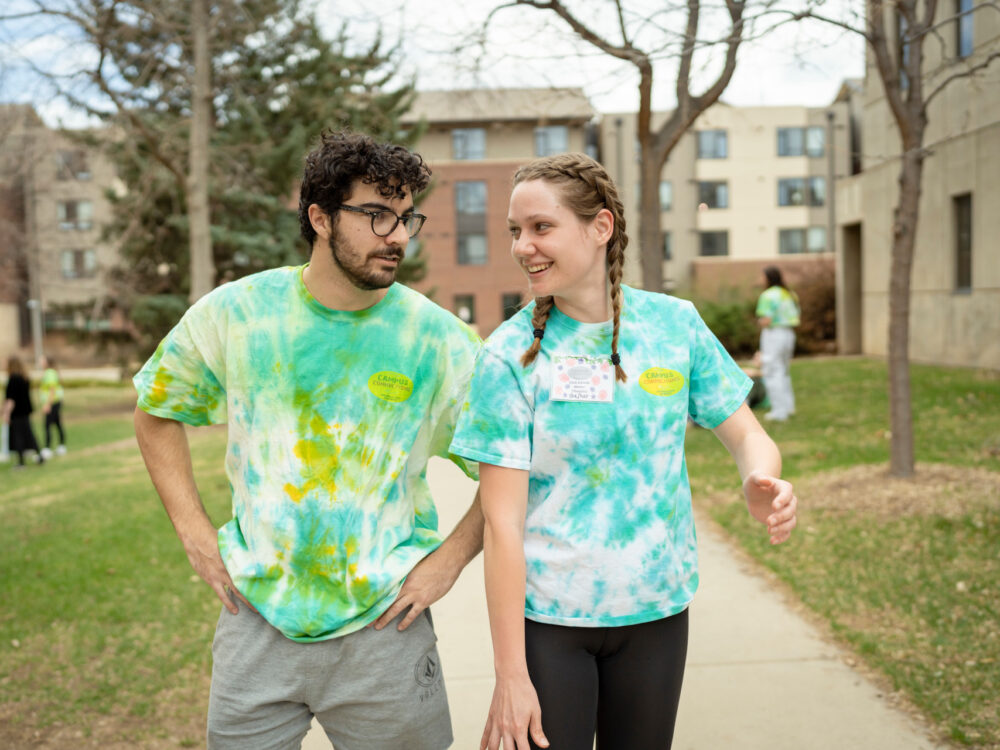 Two students in campus connections shirts walk together outside, one has hands on their hips while the other one smiles and strides