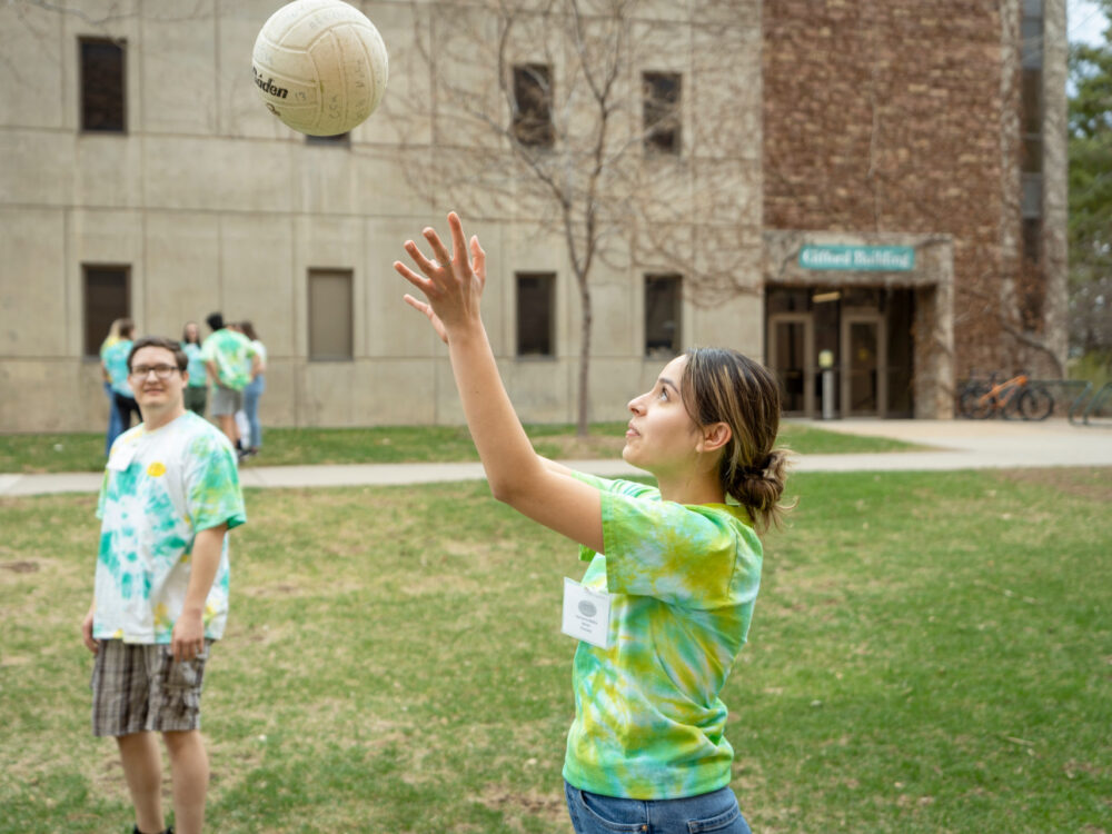 Outside, one student has their hands up prepared to set an incoming volleyball while another student watches