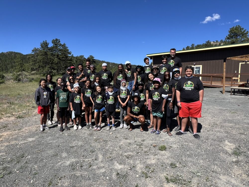 A group photo of Dandelions Rising participants, facilitators, and volunteers outside of a cabin on a clear summer day.