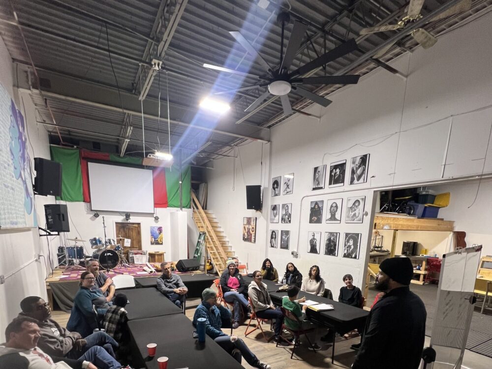 A workshop or meeting taking place in the Cultural Enrichment Center of Fort Collins. A person wearing all black with a hat stands at the front by a whiteboard, addressing a diverse group of approximately 15 attendees seated on couches and chairs around tables.