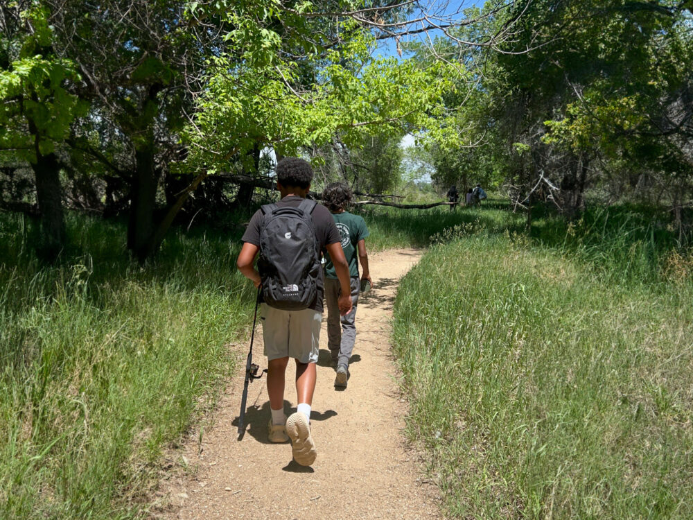 Two hikers traversing a narrow dirt trail through a verdant woodland. One person carries a fishing rod while walking ahead on the path. The trail is flanked by tall grass and creates a natural corridor beneath a canopy of bright green trees.