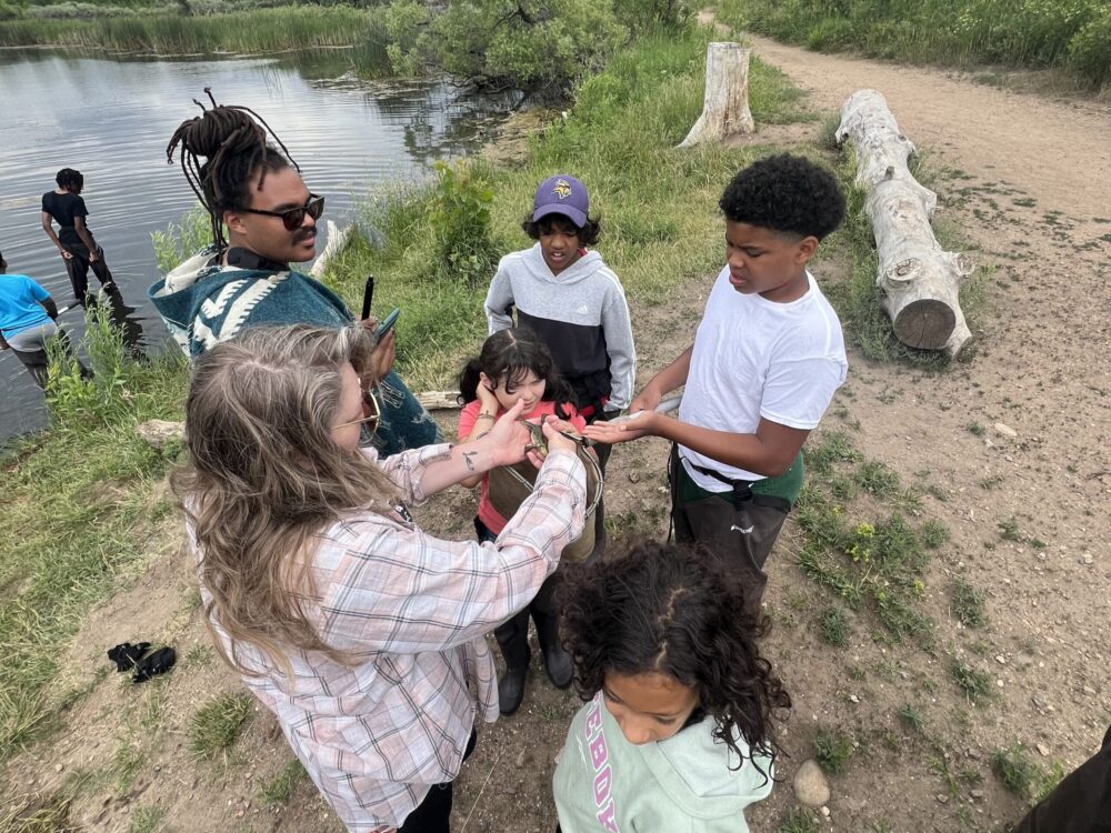 A group of Dandelions Rising participants and partners gathers on a lakeside trail, observing a frog held by one of the adults.