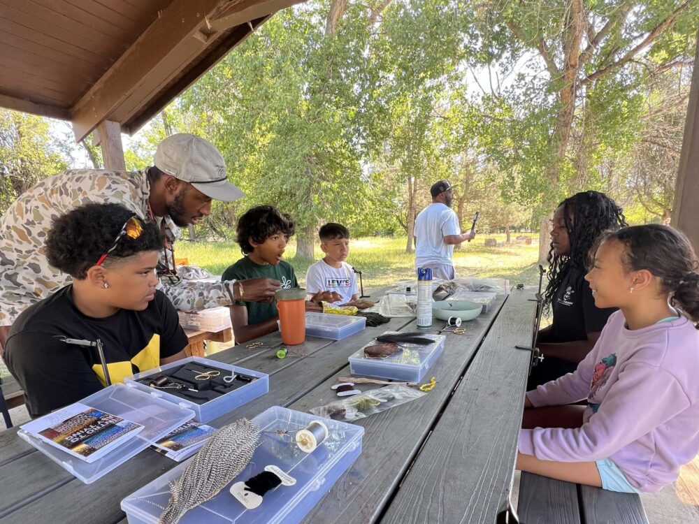 Dandelions Rising participants sit at a picnic table under a shelter, learning about fly fishing with tools and materials laid out in front of them as an instructor demonstrates