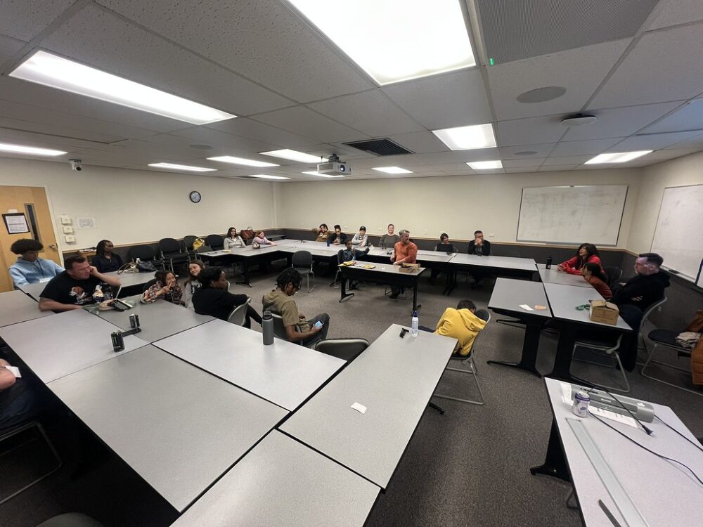 A group of Dandelions Rising participants, consisting of children and adults, engage in conversation in a classroom.