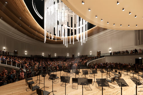 Rendering of large stage performance space with seats and musical stands on the stage and large seating area for the audience. A large crystal lighting structure hangs from the ceiling.