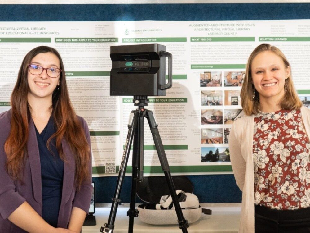 Two students stand in front of a research poster with the Matterport camera between them.