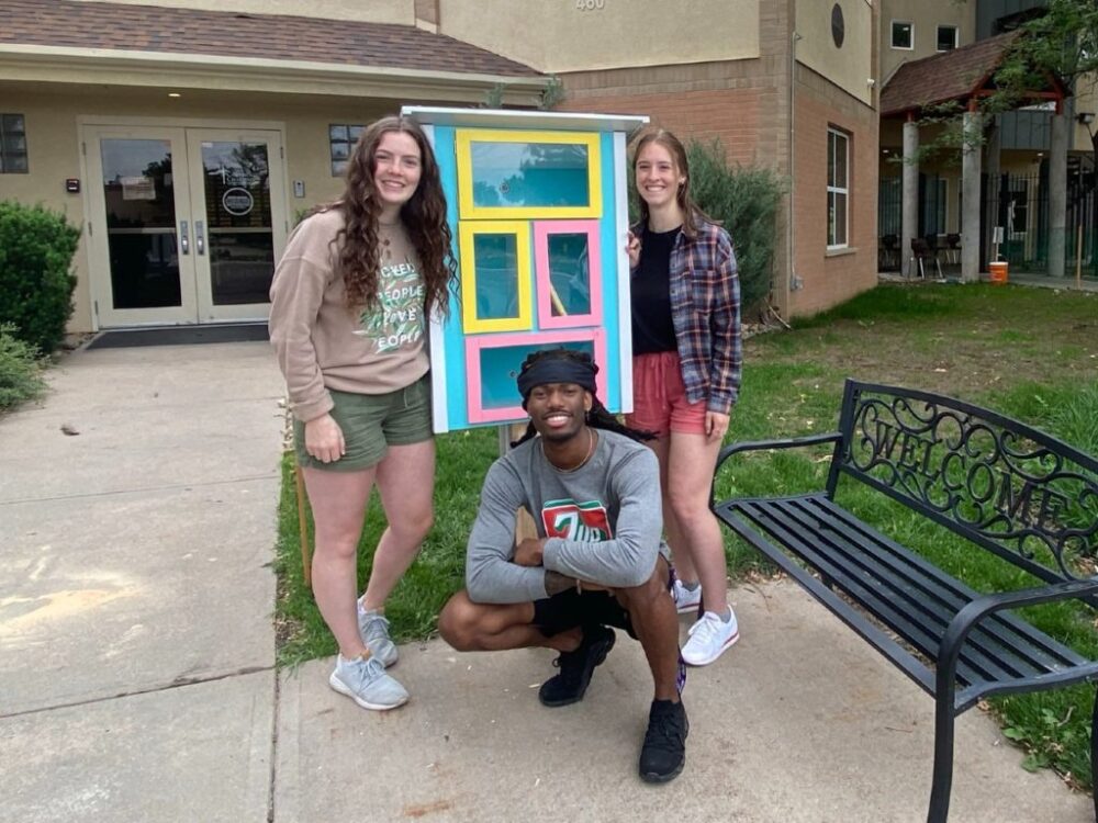 Three students stand in front of the little library they constructed in front of Catholic Charities.