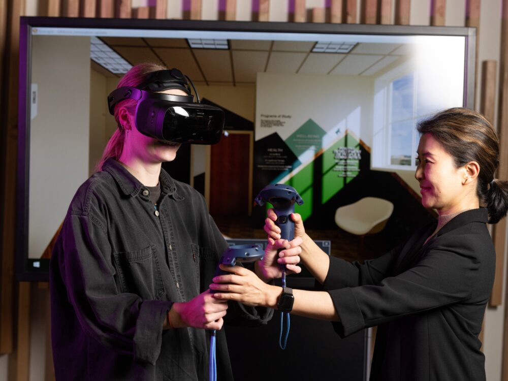 A student earning an M.S. degree in Design and Merchandising taking part in our programs and degrees wears a virtual reality headset while being assisted by a professor. The view from the headset is projected behind her.