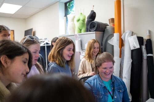 a group of students laugh with a teacher as the look at the results from the 3D body scanner in a fashion fundamantals community engagement project