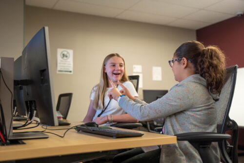 2 students sit in a computer lab, one explaining something to the other with her fingers
