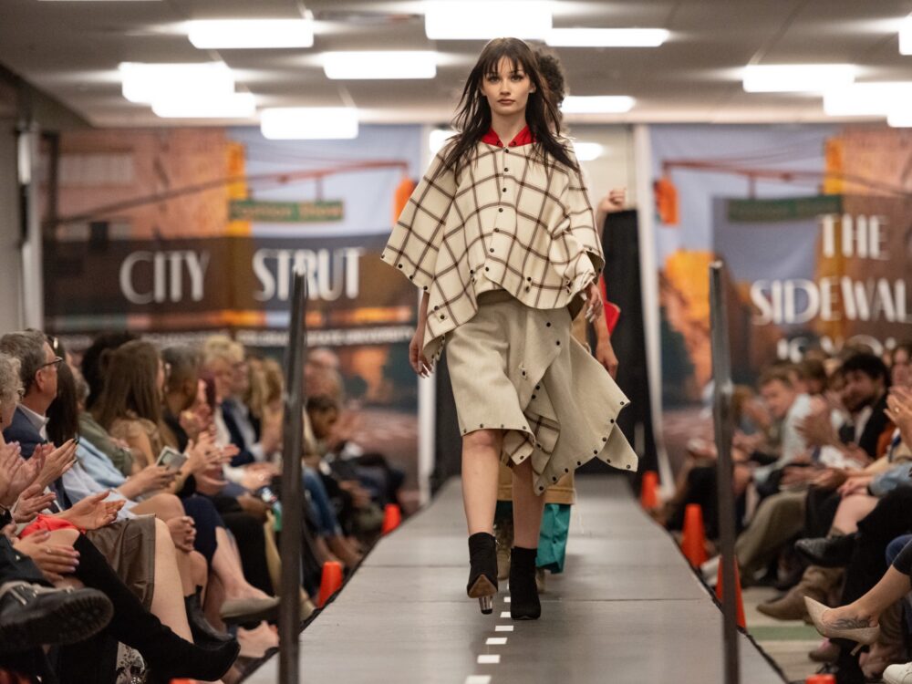 a line of models walk down the runway with a large group of audience members applauding on each side of the runway.