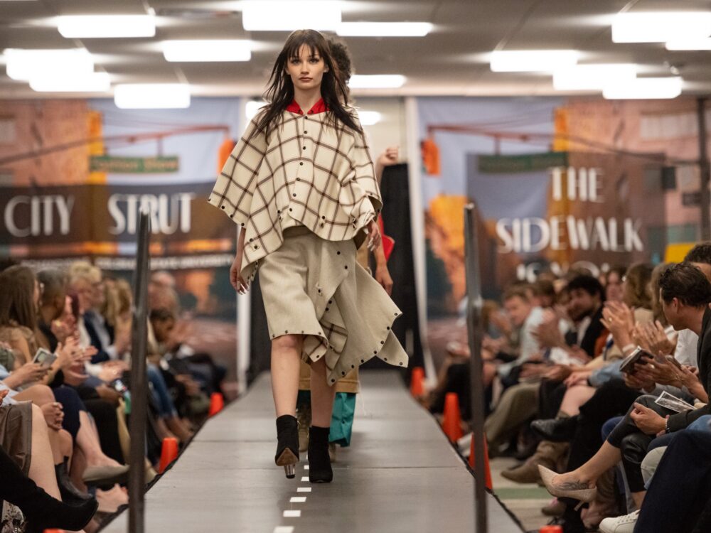 a line of models walk down the runway with a large group of audience members applauding on each side of the runway.