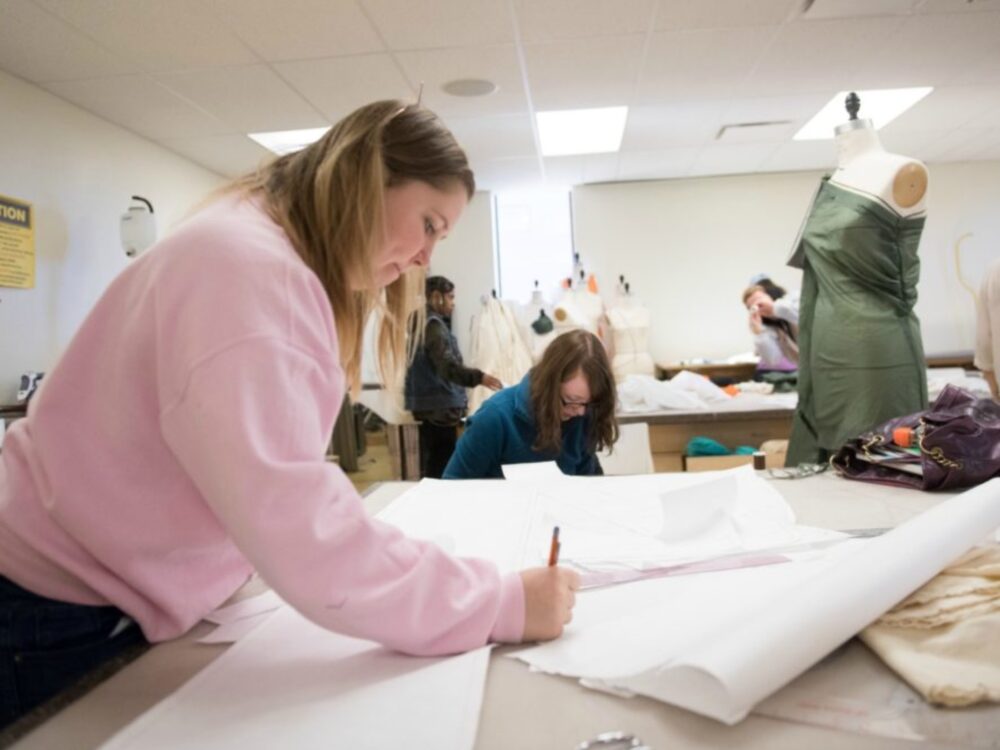 a student writes on a large piece of paper as she drafts a pattern with dress forms and other students working in the background