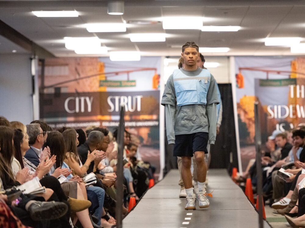 A young man walks down a catwalk with audience on each side clapping and City Strut banners in the background.