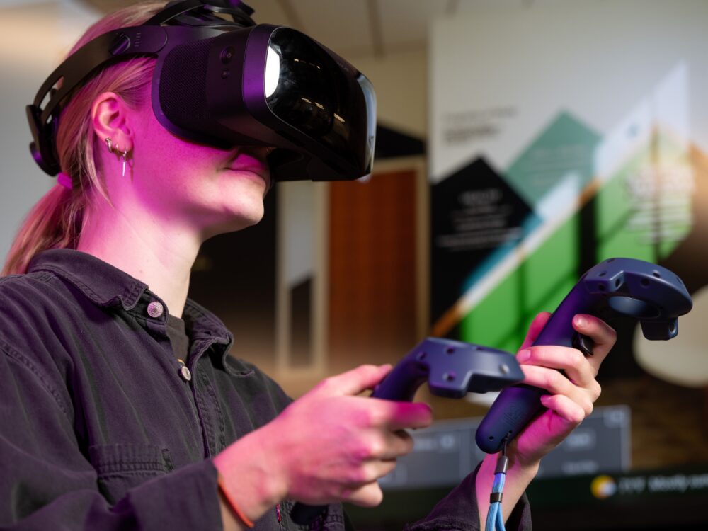 A student uses virtual reality equipment, two controllers and goggles, to view the interior of a building.
