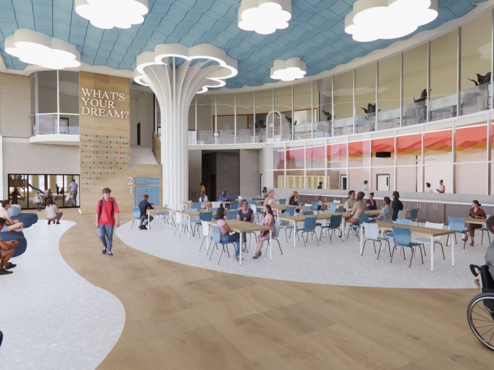A bright student commons with a cloud‑shaped ceiling, tall sculptural column, café seating, and a large wall graphic reading “WHAT’S YOUR DREAM?” beside a multi‑level staircase.