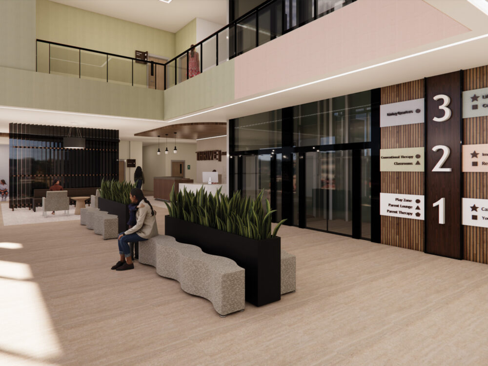 A bright community atrium with tiered seating, modern planters, a reception desk, and wall signage indicating floor levels for therapy, recreation, and café spaces.