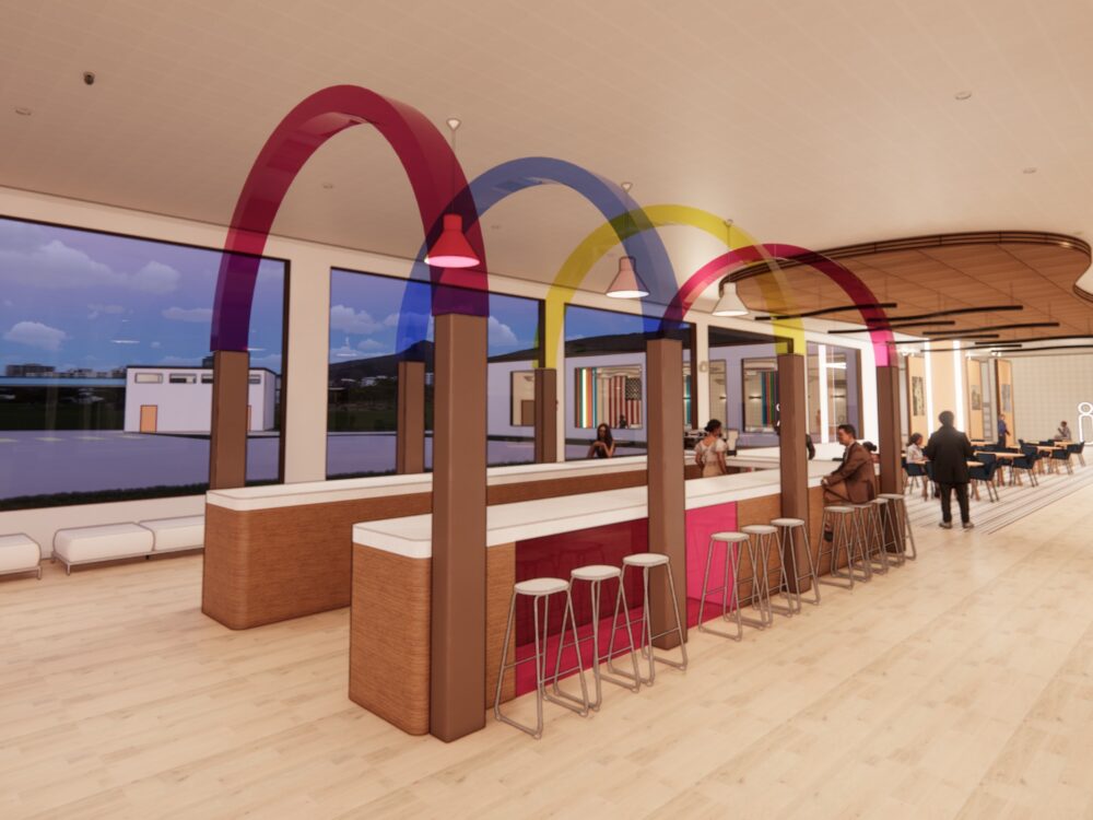 A bright café interior with long communal counters, colorful arch structures, bar stools, and large windows lining the space.