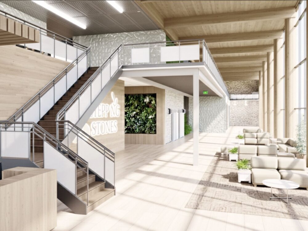 A bright lobby with tall windows, light wood finishes, a central staircase leading to an upper walkway, lounge seating along the window wall, and a “STEPPING STONES” sign set into a living‑plant feature wall