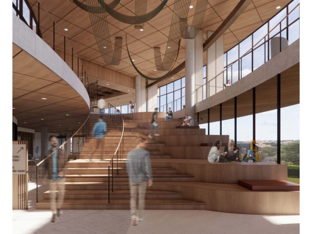 Interior architectural rendering of a large open stair/lounge area with tiered wooden seating, floor‑to‑ceiling windows, and a curved upper walkway. Sculptural hanging elements decorate the ceiling. A directional sign indicates access to “2” and “Library, Study Hall, Community Services.” People sit, walk, and gather on the steps, with views of greenery visible through the windows.