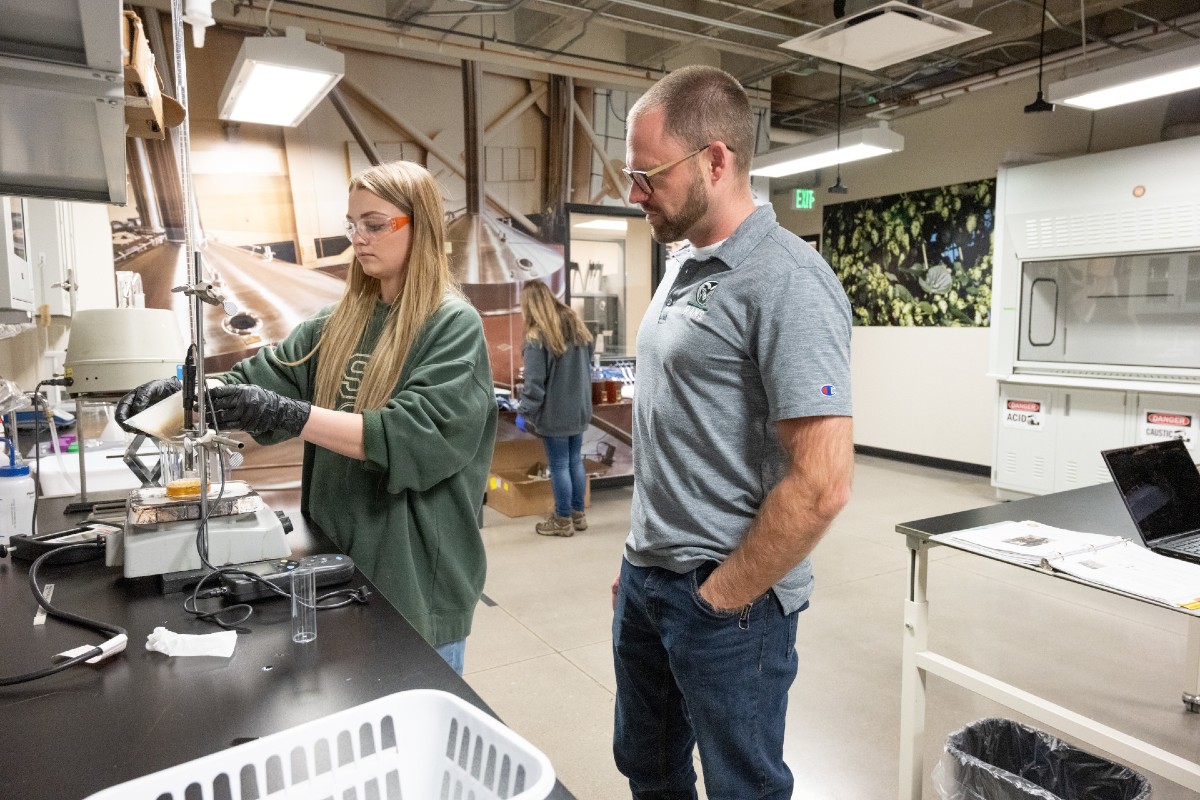 A student wearing gloves and eye protection works at a lab counter as an instructor looks over her shoulder