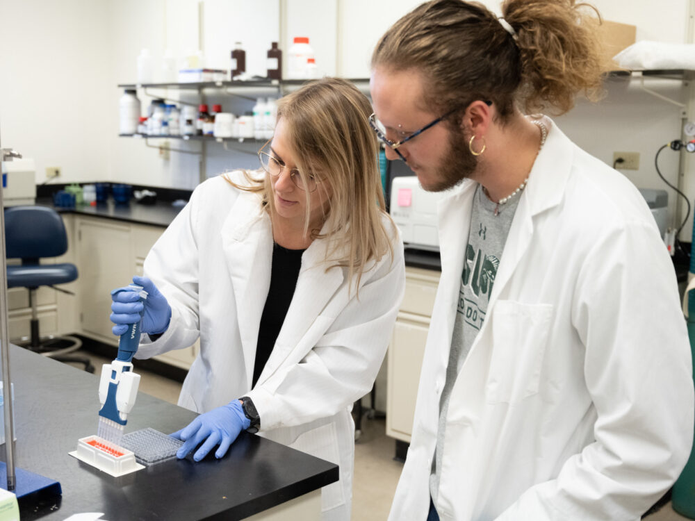 A researcher in a white lab coat demonstrates in a lab to a student in a white lab coat