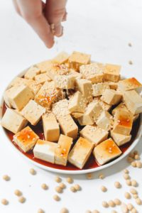 Cubed, browned, tofu on a plate being sprinkled with sesame seeds.