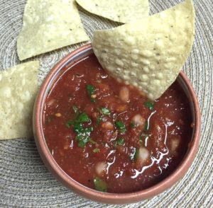 A bowl of salsa containing cilantro with corn chips laying around
