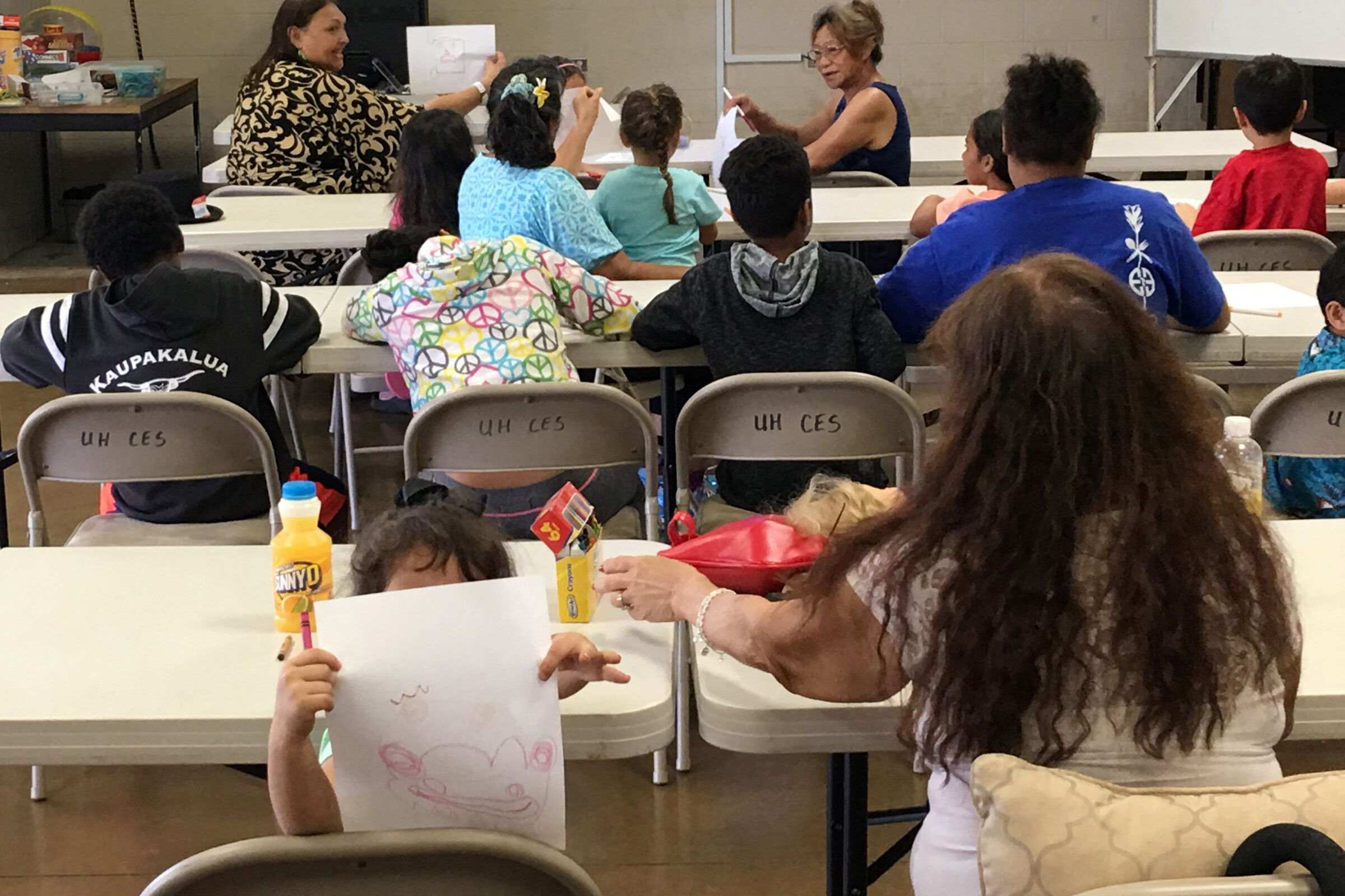 Children and adults sitting at tables drawing - in the forefront a small child holds up their drawing