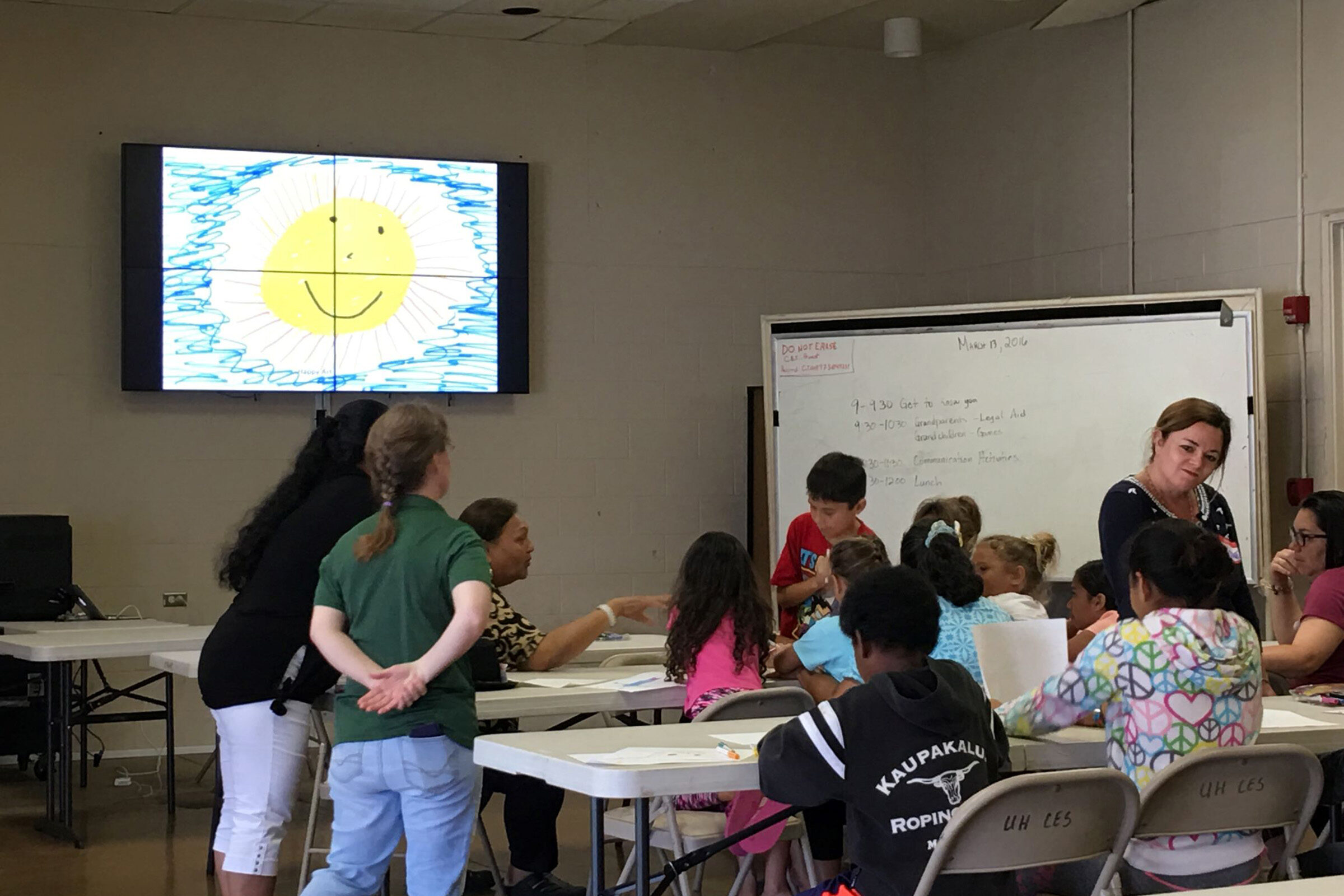 Room with kids and adults sitting at tables drawing on paper with an illustration of a sun projecting on the screen at the front of the room