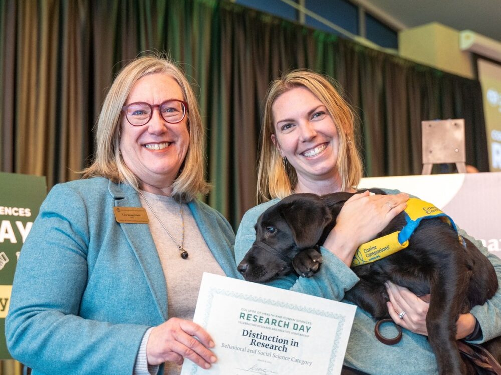 csu college of health and human sciences dean lise youngblade presenting research day award to kate miller who is holding a black lab puppy service dog in training.