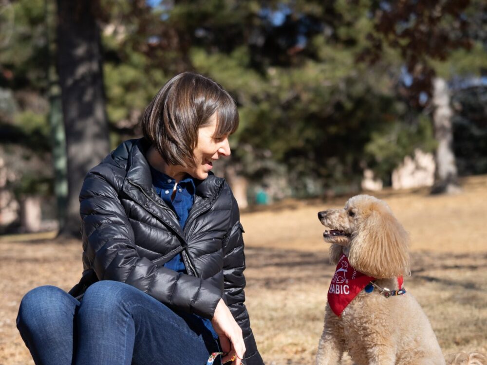 habic volunteer abby and her dog mischa, a medium-sized poodle with sand-colored fur, sitting in a park looking at each other and smiling