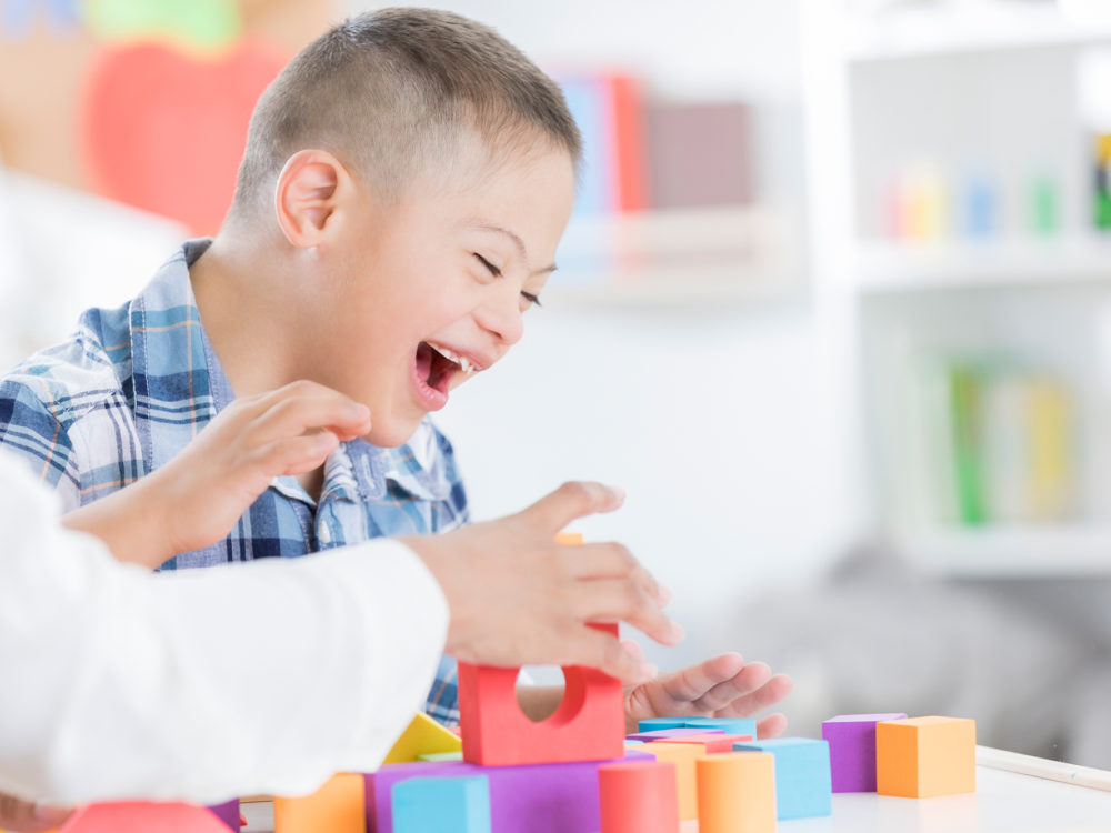 Cheerful preschool age boy enjoys playing with blocks with his teacher. Only the teachers arms are seen in the photo.
