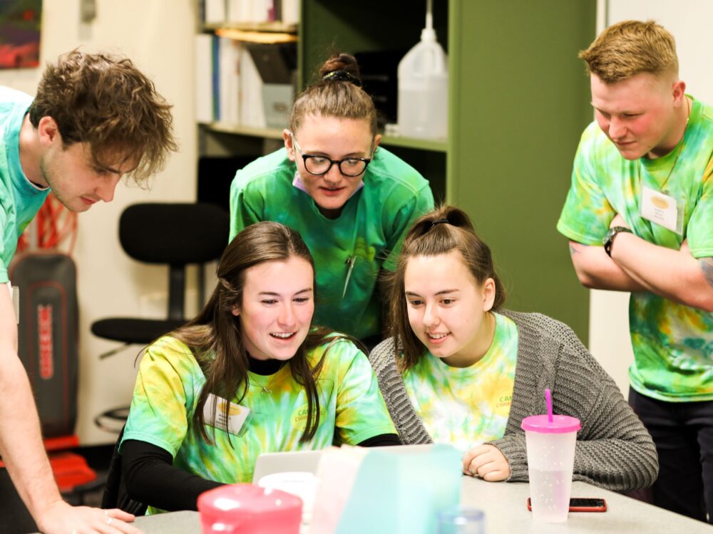 Group of students wearing green, yellow, and white tie dye shirts collaborate around a laptop
