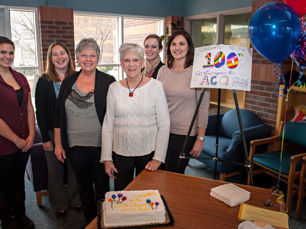 Group of 5 smile with sign that says `100` and a cake in front of them
