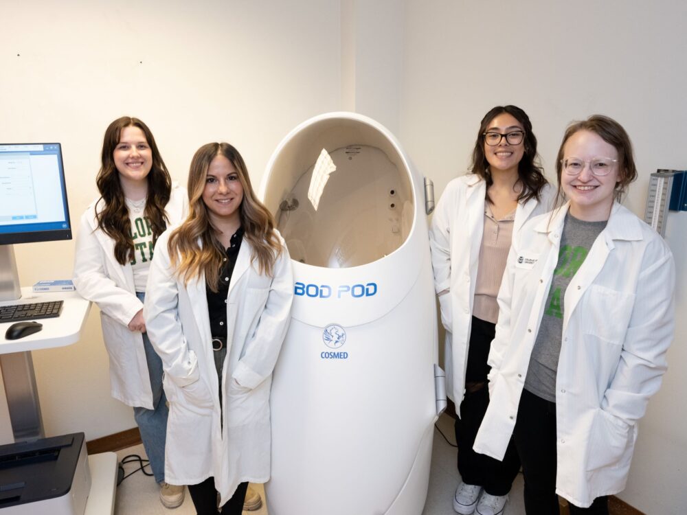 Four individuals wear lab coats and stand in front of human sized pod.