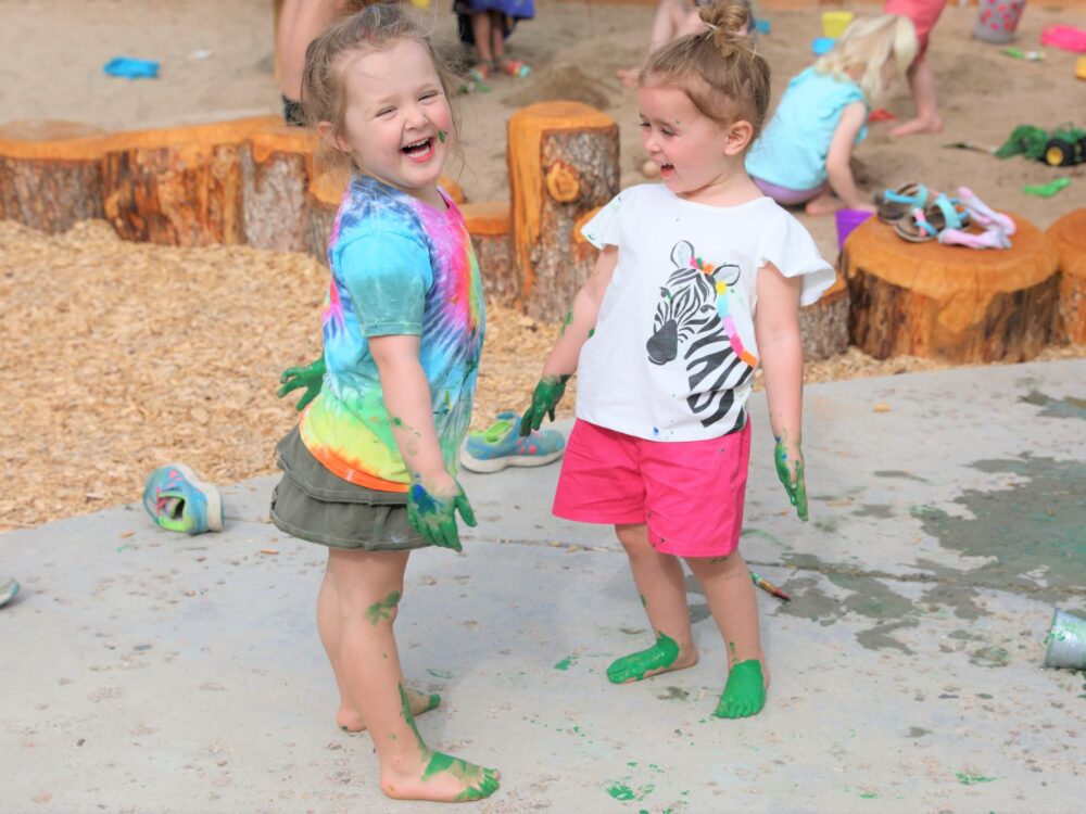 Two girls smile with green paint on their arms and legs.