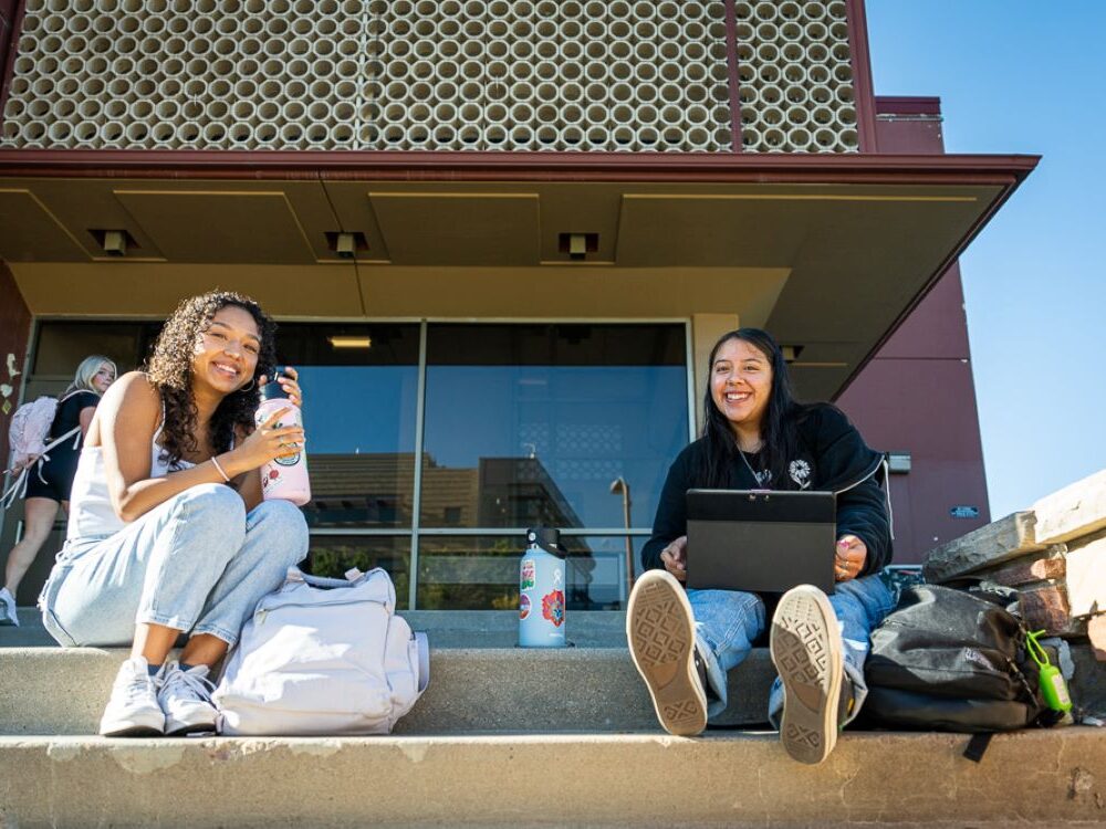 Two students sit on steps and smile